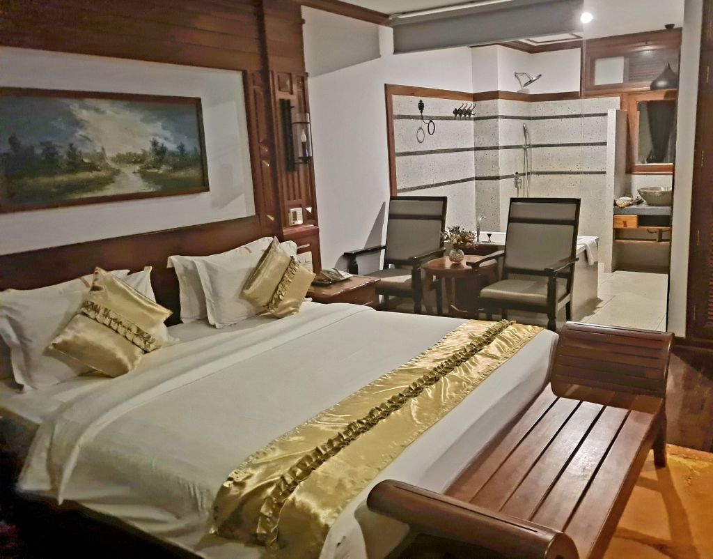 A luxurious bedroom featuring a large bed with decorative pillows, a wooden bench, and elegant bedding accented with gold. To the right, there is a seating area with two chairs and a wooden table, along with a modern bathroom visible in the background.