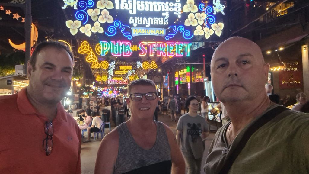 Three men posing for a selfie at night on Pub Street, showcasing vibrant neon lights and a bustling street scene in the background.