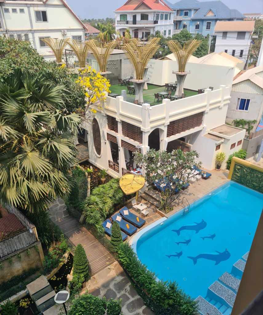 Aerial view of a hotel courtyard featuring a swimming pool with dolphin designs, surrounded by lounge chairs, tropical plants, and a patio area with tables under palm-like structures.