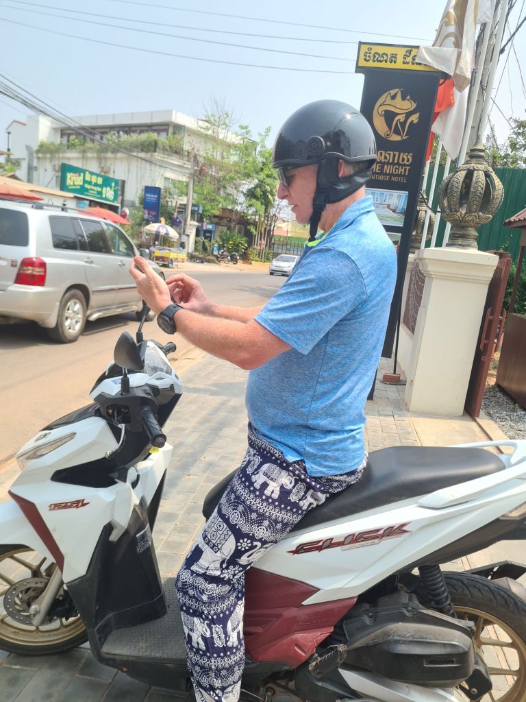 A person wearing a helmet is sitting on a motorcycle, checking their phone, dressed in a blue shirt and patterned pants, on a street with various buildings and vehicles in the background.