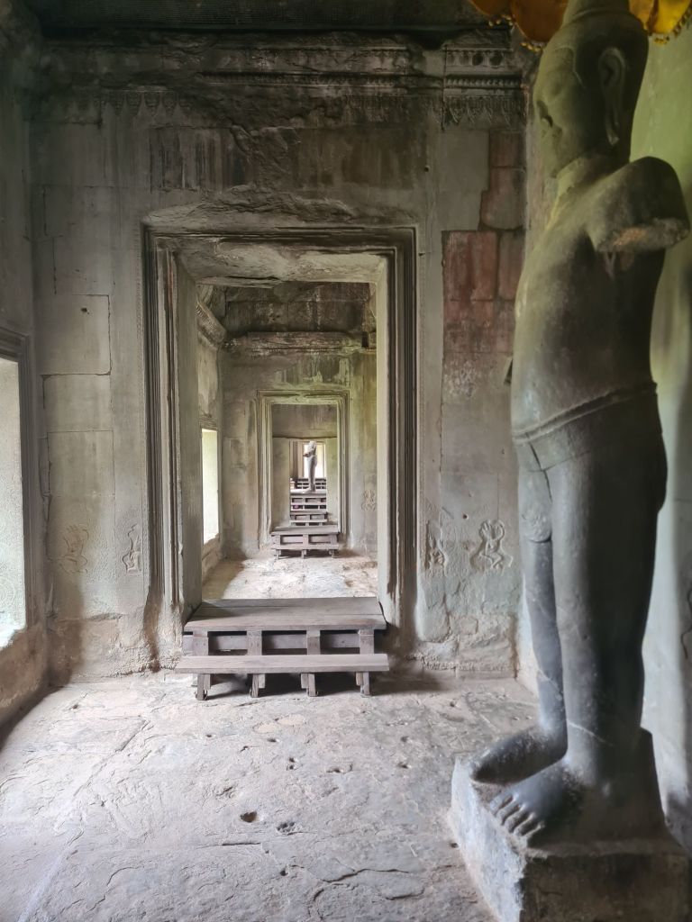 An interior view of an ancient stone structure featuring a long hallway with multiple doorways, stone benches, and a prominent statue of a figure on the right.