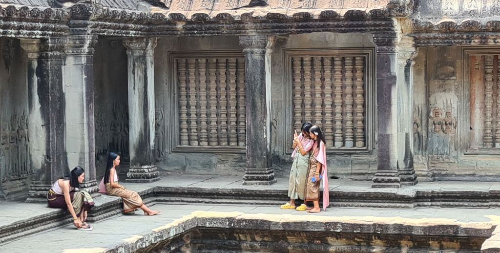 Four young girls dressed in traditional attire are gathered in an ancient temple setting, with two girls sitting on steps, one girl adjusting her clothing, and two girls standing closely together, one looking at a phone.