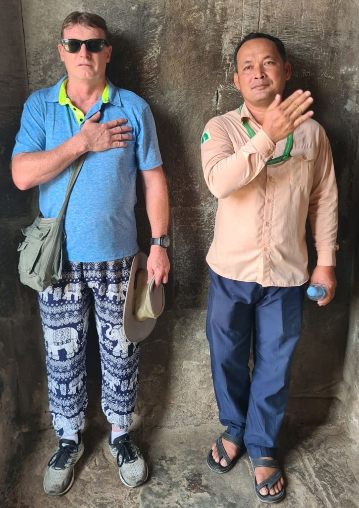 Two men standing side by side against a stone wall; one wearing sunglasses and a blue shirt with patterned pants, and the other wearing a beige shirt and dark pants, both with hands over their hearts.
