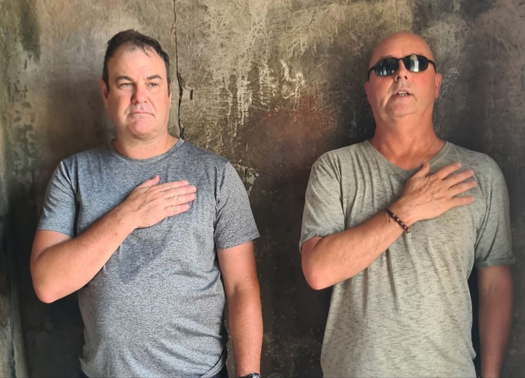 Two men standing side by side with their hands on their hearts, wearing gray t-shirts, in front of a textured wall.