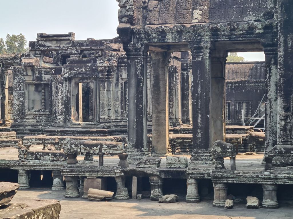 Ruins of an ancient stone structure with weathered columns and intricate carvings, surrounded by debris and overgrown vegetation.