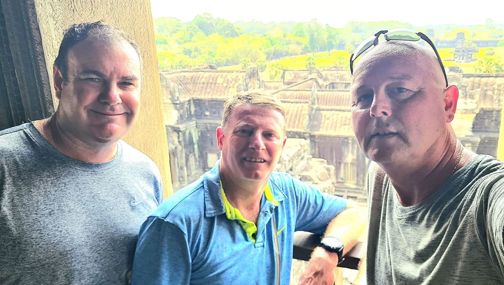 Three men posing for a selfie with ancient temple ruins in the background, greenery visible in the distance.