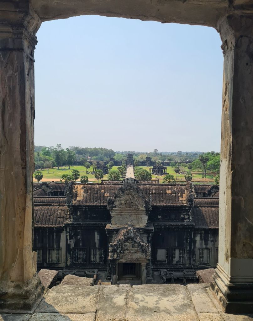 View from a stone window frame overlooking ancient temple architecture and lush green landscapes in Angkor Wat, Cambodia, under a clear blue sky.