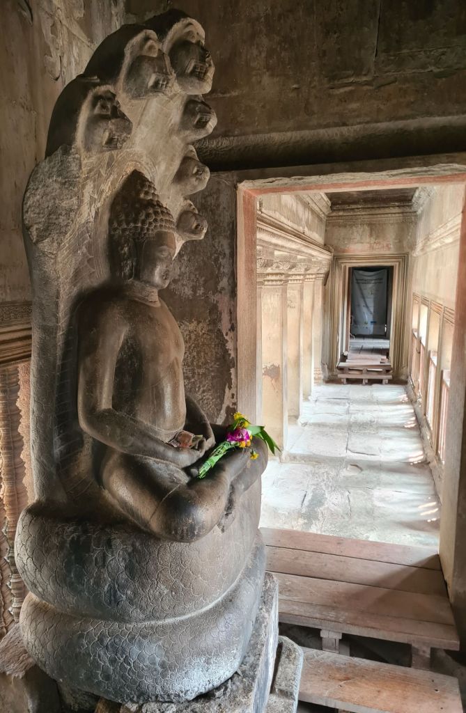 A stone Buddha statue seated in a meditative pose, adorned with a garland, located in a historic temple corridor, with intricate carvings and natural light illuminating the space.