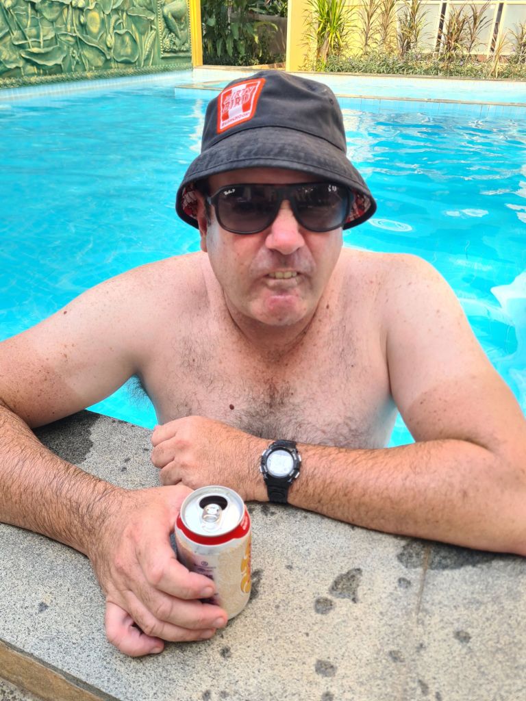 A man with sunglasses and a bucket hat sitting by the edge of a swimming pool, holding a can of beer.