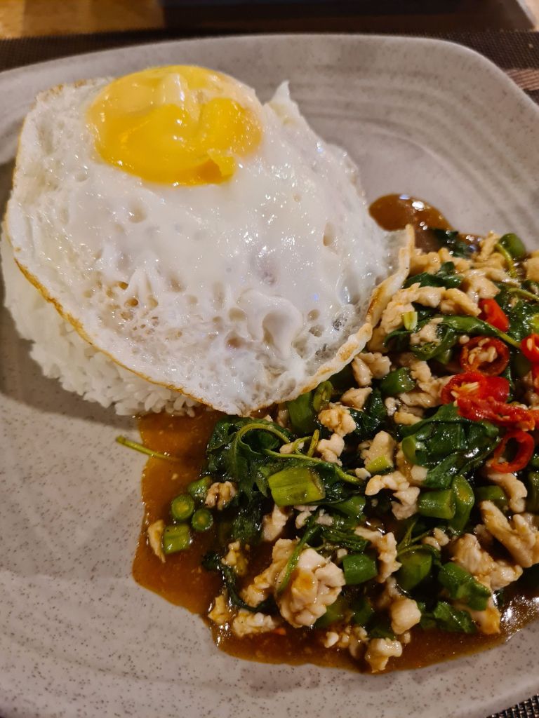 A plate of rice topped with a fried egg and a serving of stir-fried chicken with basil, green onions, and chili peppers.