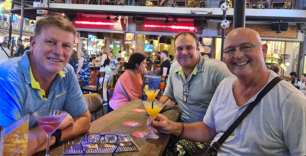 Three men smiling and enjoying drinks at a bar, with colorful cocktails on the table and a lively background.