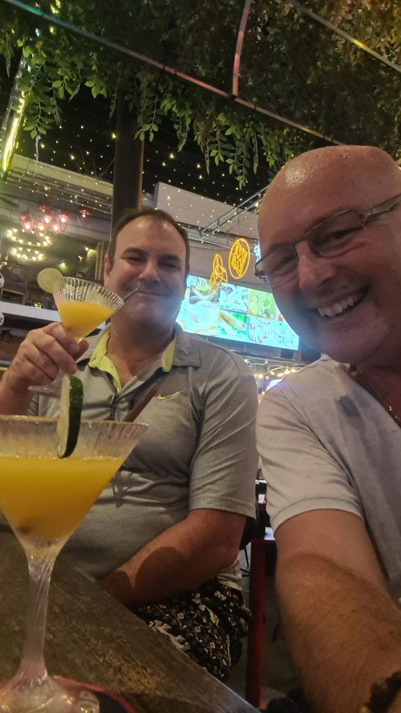 Two men smiling while sitting at a table with colorful cocktails in a lively bar setting, decorated with lights and greenery.