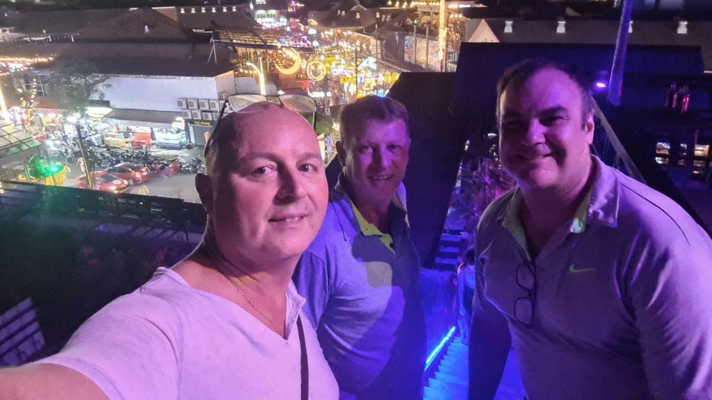 Three men posing for a selfie at a rooftop location with a vibrant cityscape in the background, featuring colorful lights and busy streets.