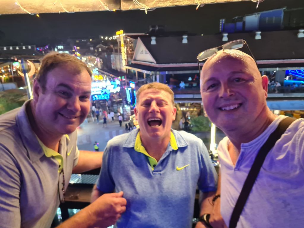 Three men smiling and posing for a selfie at night, with colorful lights and a bustling scene in the background.