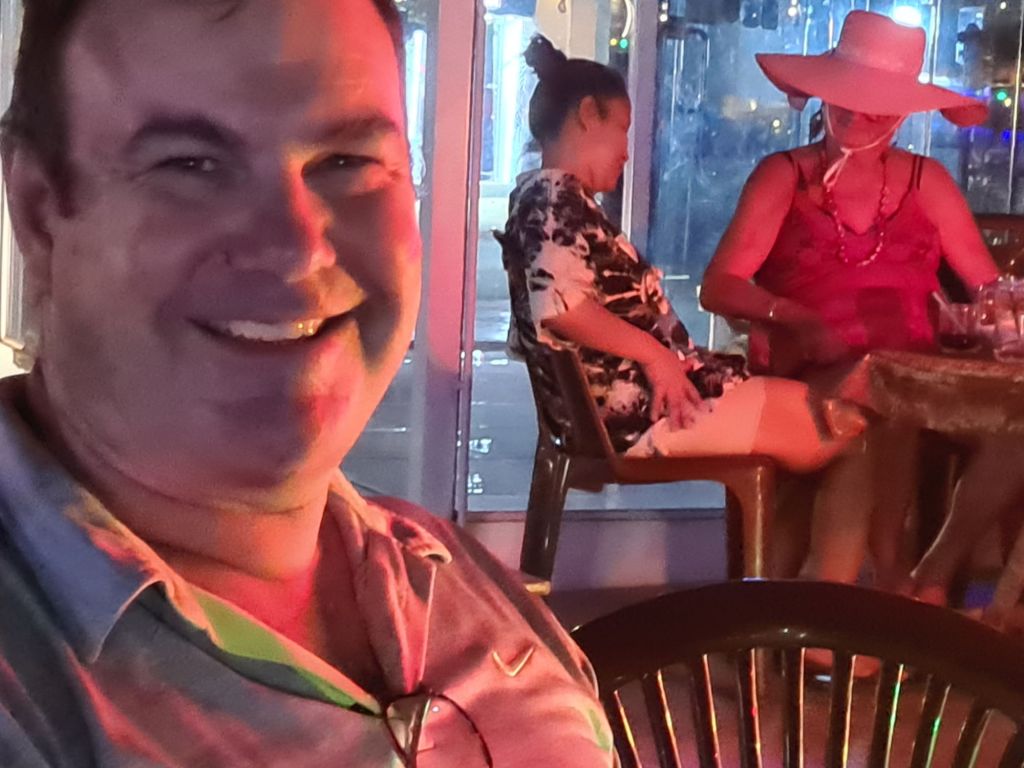 A smiling man in a casual shirt sits in a dimly lit setting, with colorful lighting. In the background, two women are seated, one wearing a floral outfit and another in a large hat.