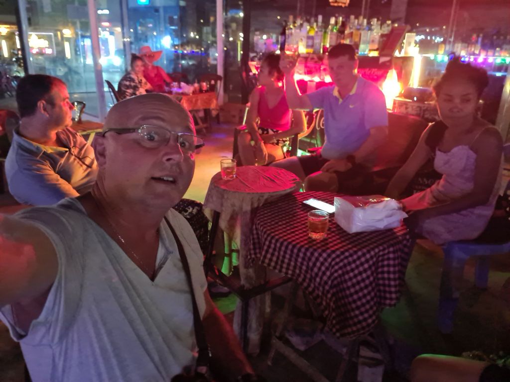 A group of five people socializing at a bar in a lively setting with colorful lighting. One person is taking a selfie, while others are seated at a table with drinks.