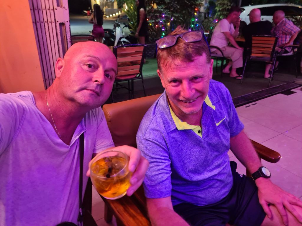 Two men sitting together in a bar, one holding a drink, with a lively atmosphere in the background.