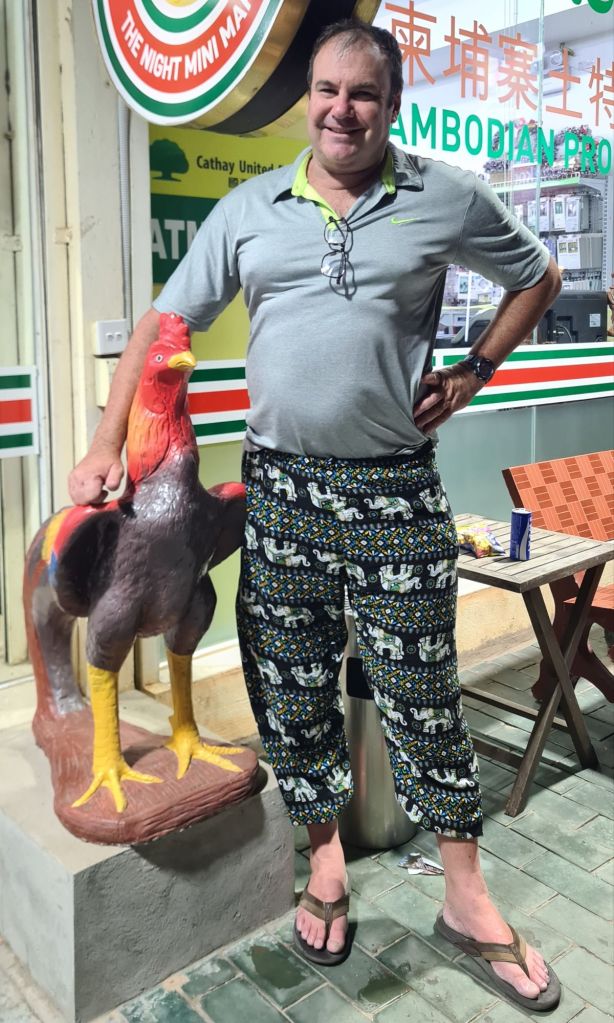 A man standing next to a colorful rooster statue, wearing a grey shirt and patterned pants, with flip-flops on a tiled floor.