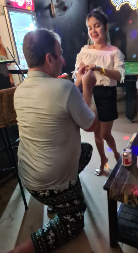 A man kneeling on one knee is proposing to a woman in a casual setting, with a joyful expression on her face. The background features soft lighting and colorful decorations.