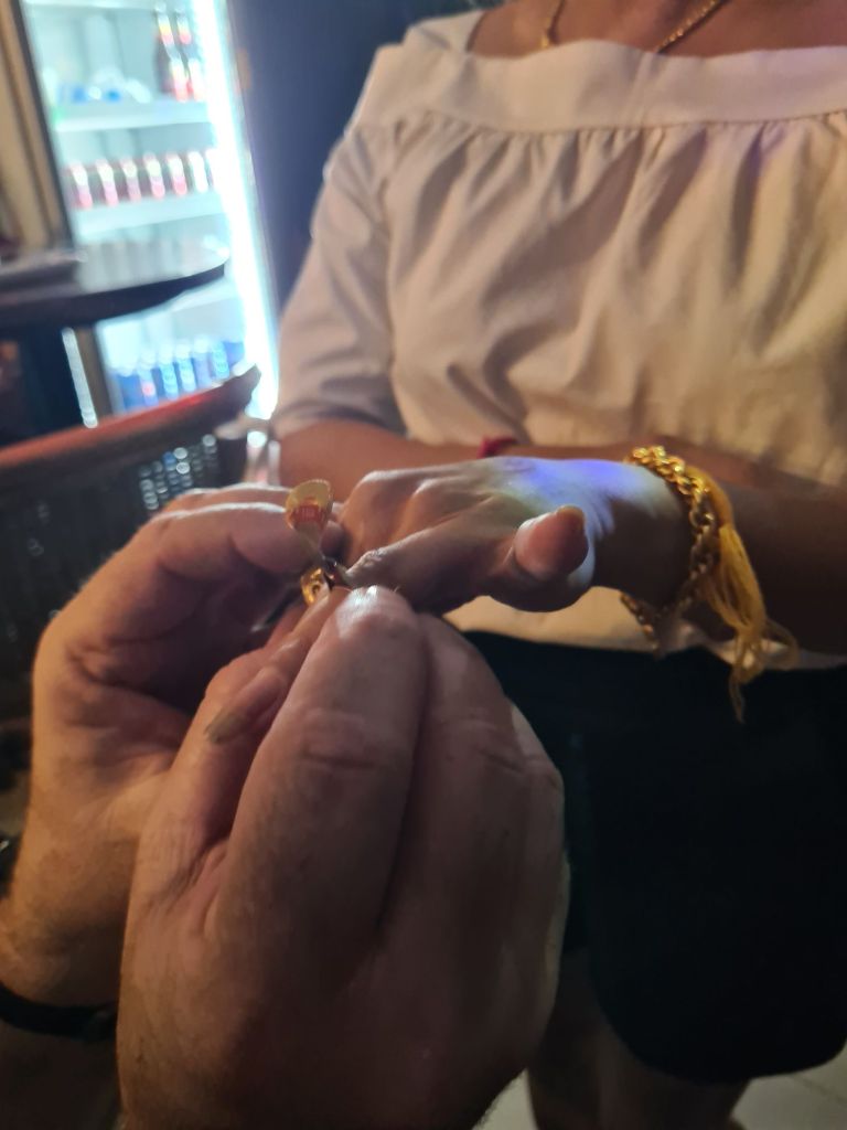 A close-up of a person’s hand receiving a ring from another person, with jewelry and a brightly lit background.
