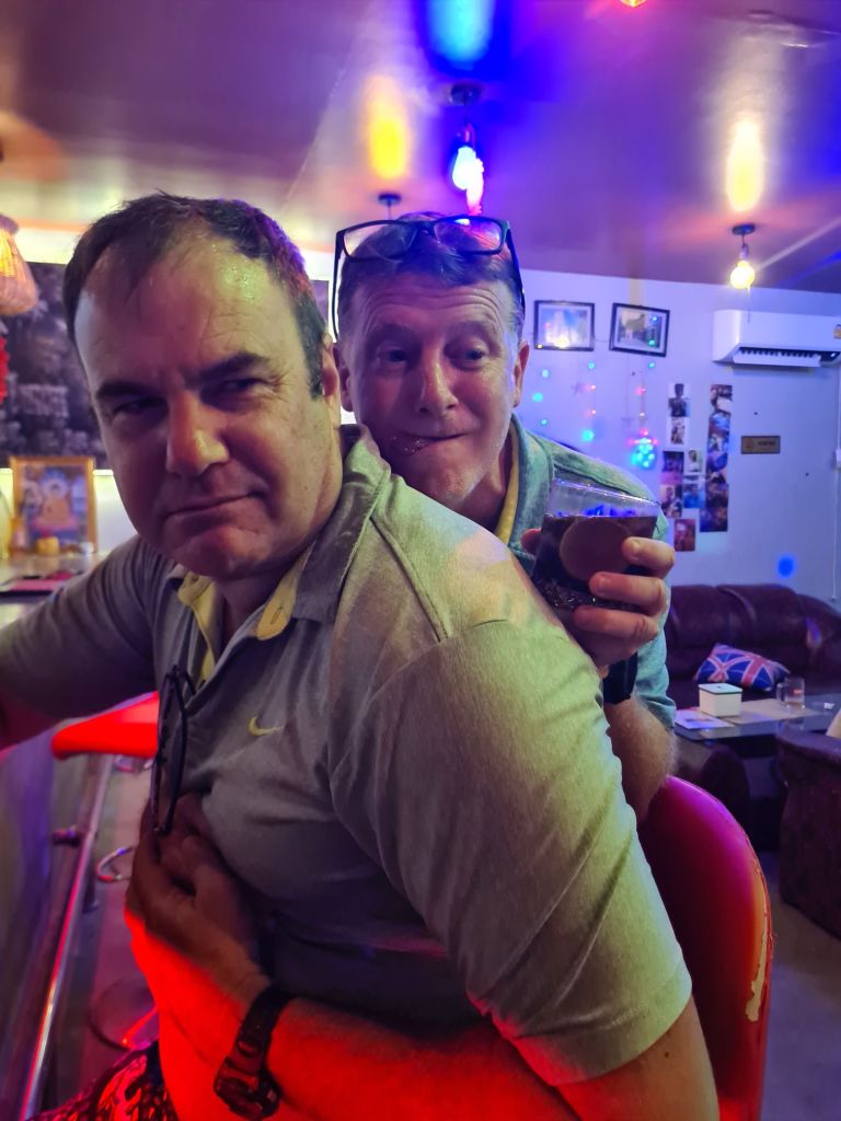 Two men in a colorful bar setting, one sitting and looking serious while the other playfully leans in behind him, holding a drink.