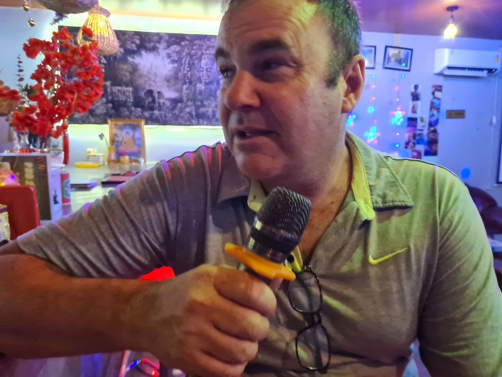 A man holding a microphone while sitting in a colorful, lively setting with decorative items and a backdrop featuring artwork.