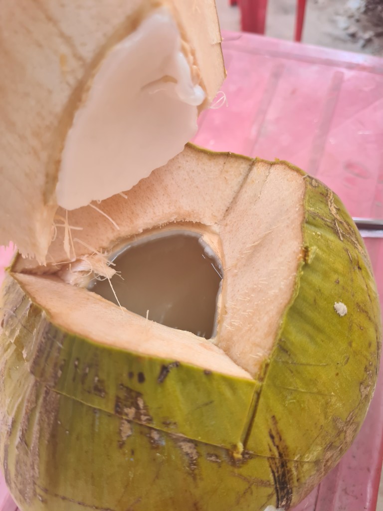 A freshly opened green coconut with soft white flesh exposed and liquid inside, placed on a pink table.