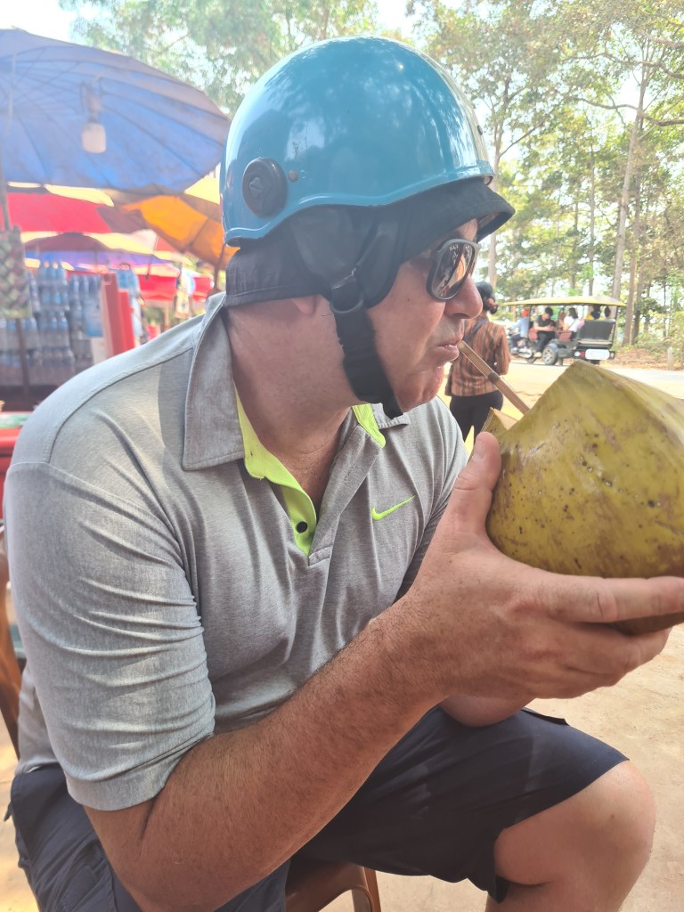 A man wearing a blue helmet and sunglasses sitting outdoors, sipping from a coconut through a straw, with colorful umbrellas in the background.