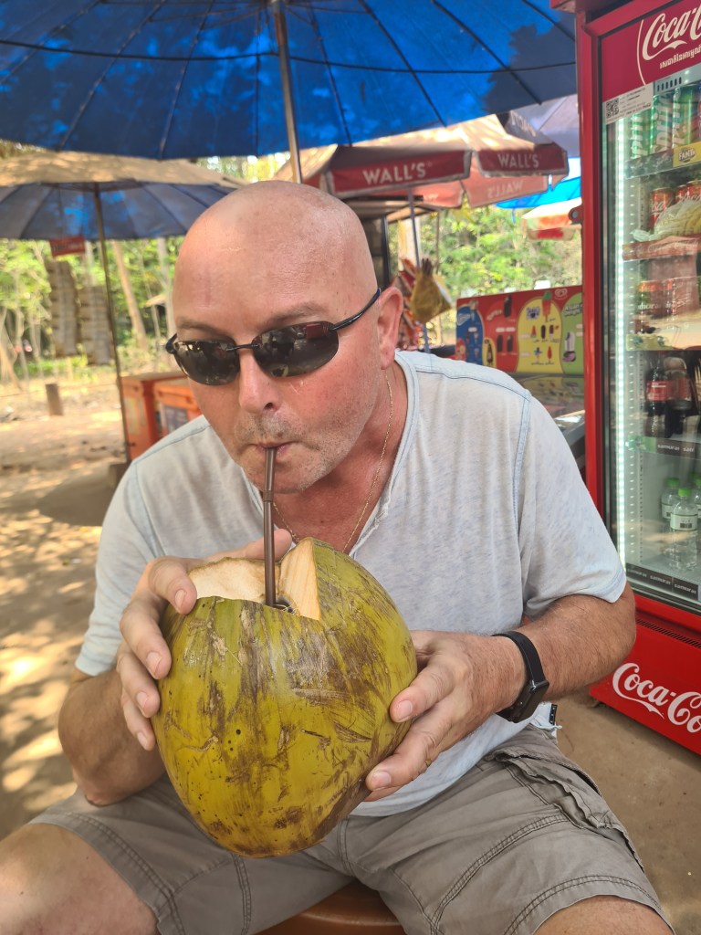 A person with a shaved head and sunglasses drinking coconut water through a straw from a fresh coconut while sitting under an umbrella at an outdoor vendor stall.