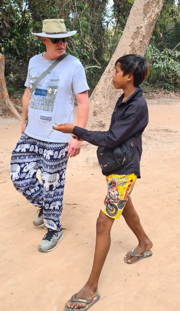 An adult man wearing a gray t-shirt and patterned pants walks alongside a young boy dressed in a black jacket and bright shorts, engaged in conversation on a sandy path surrounded by trees.