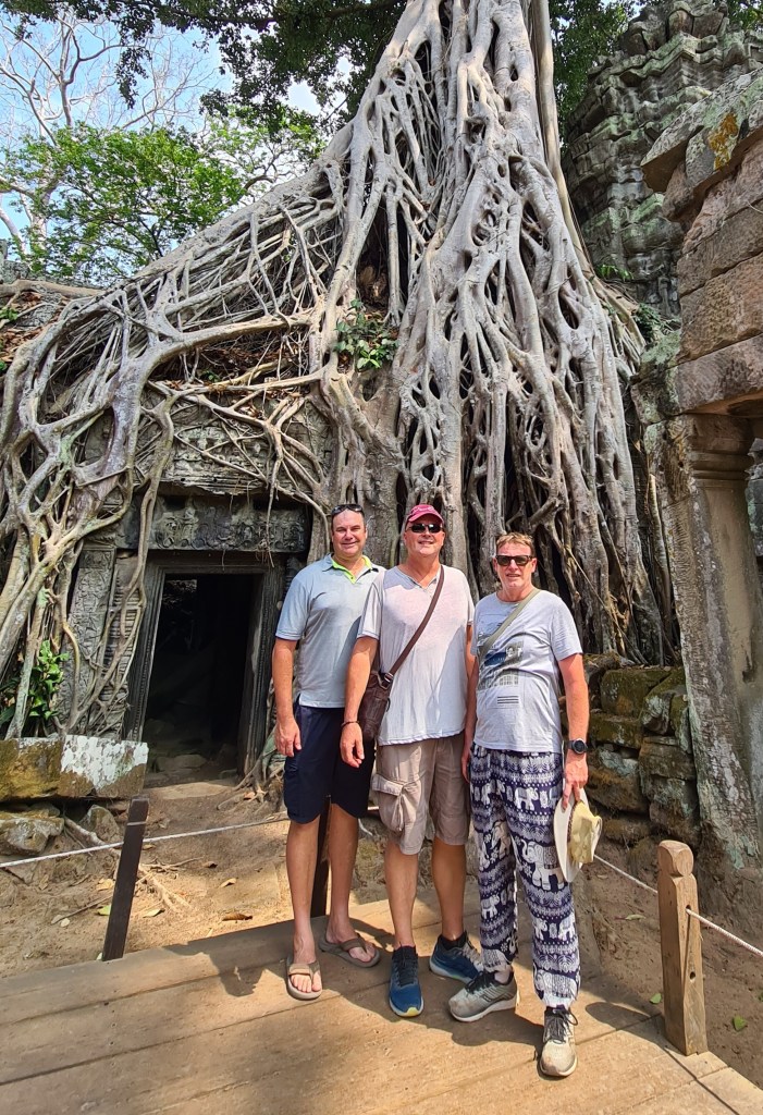 Three people posing in front of a large tree with exposed roots, next to a stone structure, in a natural setting.