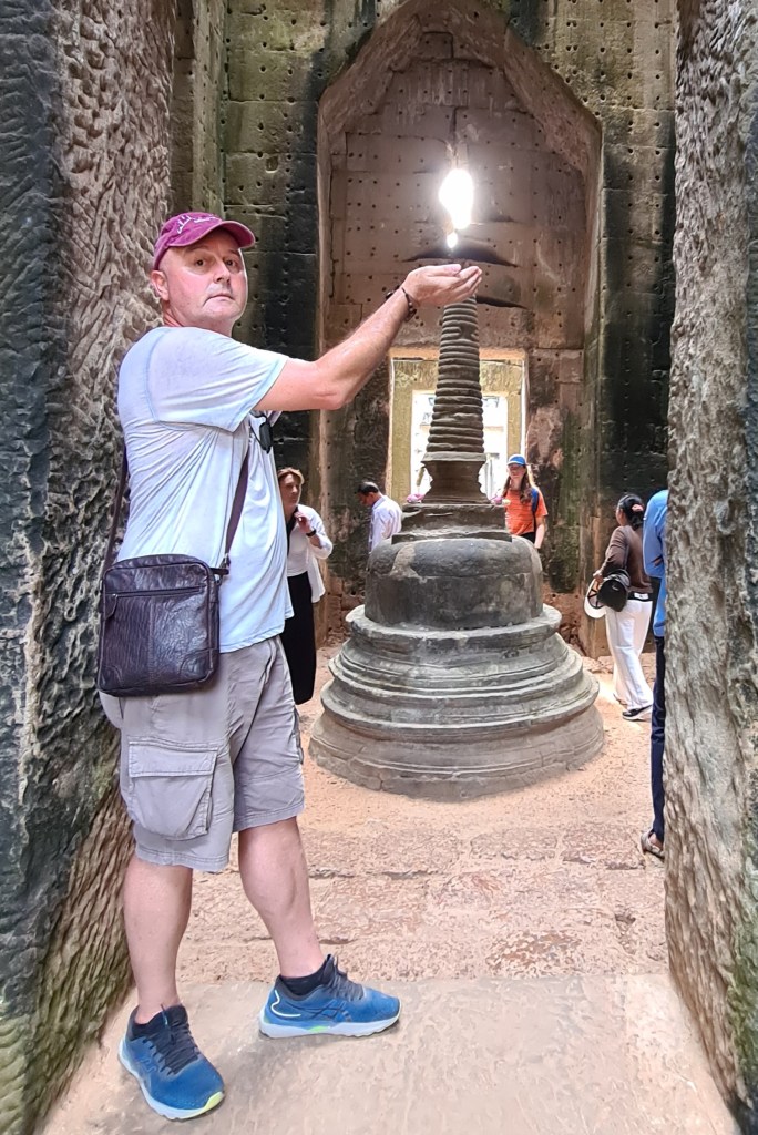 A man posing inside a historical temple, holding his hand up as if supporting a glowing light above a stone structure. The temple features ancient stone walls and several tourists in the background.
