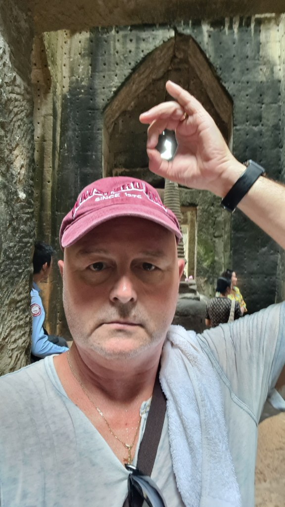 A person wearing a maroon cap is taking a selfie inside a stone structure, holding a hand in a gesture above their head. The background shows other visitors and stone walls.