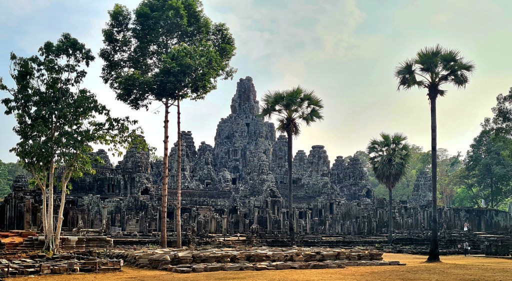 Ancient temple ruins surrounded by lush trees and palm trees, with a misty sky in the background.