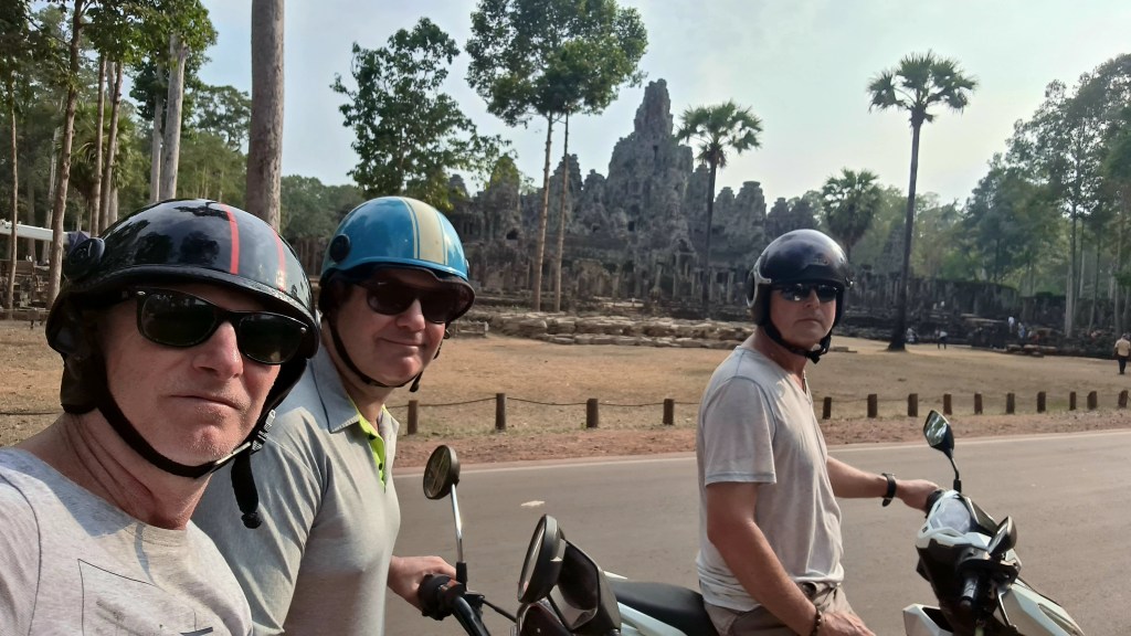 Three men wearing helmets pose for a selfie while on scooters, with ancient temple ruins visible in the background surrounded by trees.