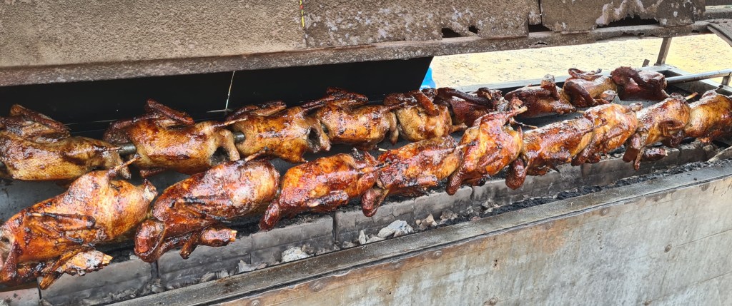 A row of whole chickens being roasted on a spit over an open flame, with a shiny, golden-brown skin.