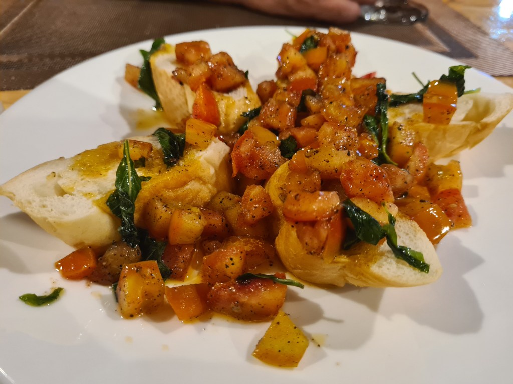 A close-up of sliced bread topped with a mixture of diced tomatoes, bell peppers, and basil, drizzled with olive oil and seasoned with black pepper, served on a white plate.
