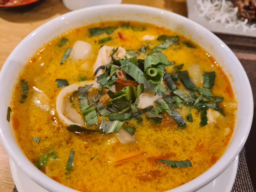 A bowl of rich and aromatic soup with yellow broth, featuring sliced vegetables and herbs on top.