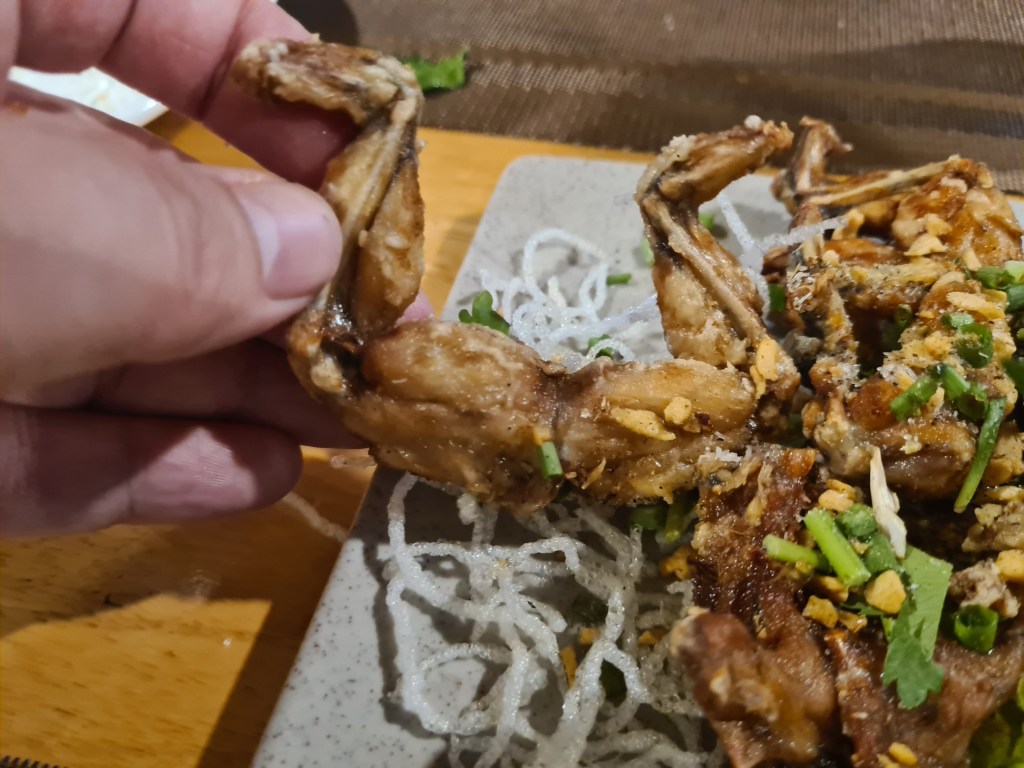 A person holding a fried frog leg, with a plate of garnished fried frog legs in the background.