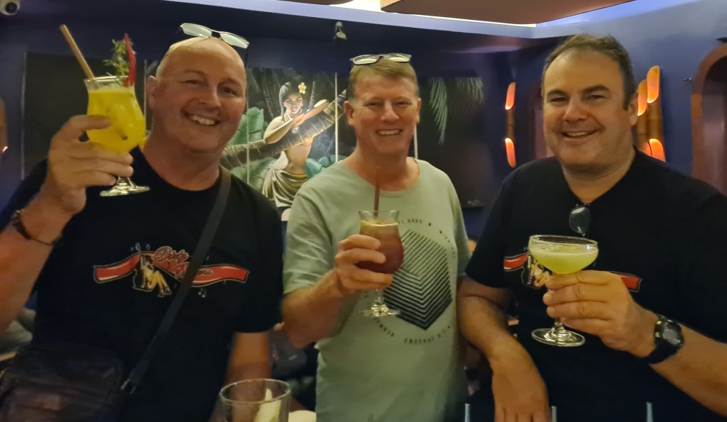 Three men smiling and holding colorful cocktails in a bar setting.