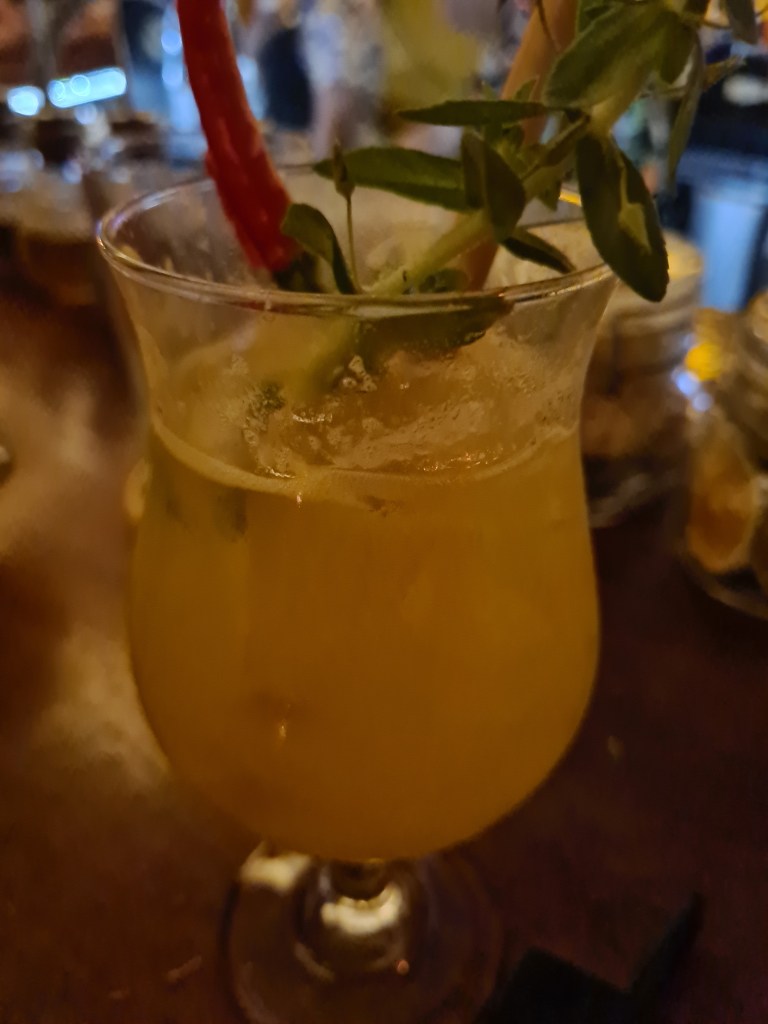Close-up of a cocktail glass containing a yellow drink, garnished with a red chili pepper and fresh herbs, set on a wooden surface.