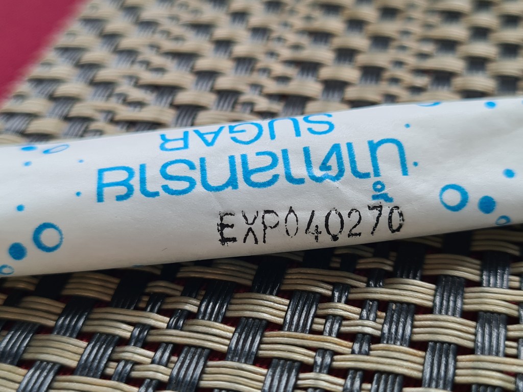 A close-up of a wrapped sugar packet with the text 'Ultimate Sugar' and an expiration code 'EXP040270' printed on it, placed on a woven surface.