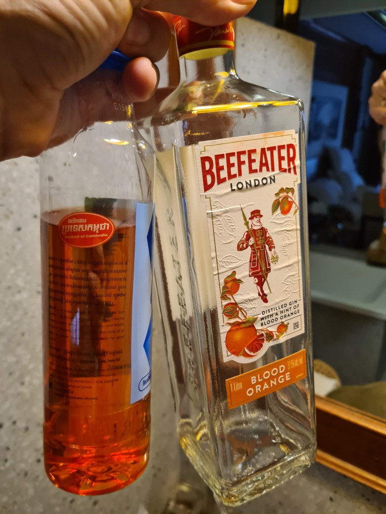 A hand holding a bottle of Beefeater Blood Orange gin next to a bottle of a reddish-orange beverage in a kitchen setting.