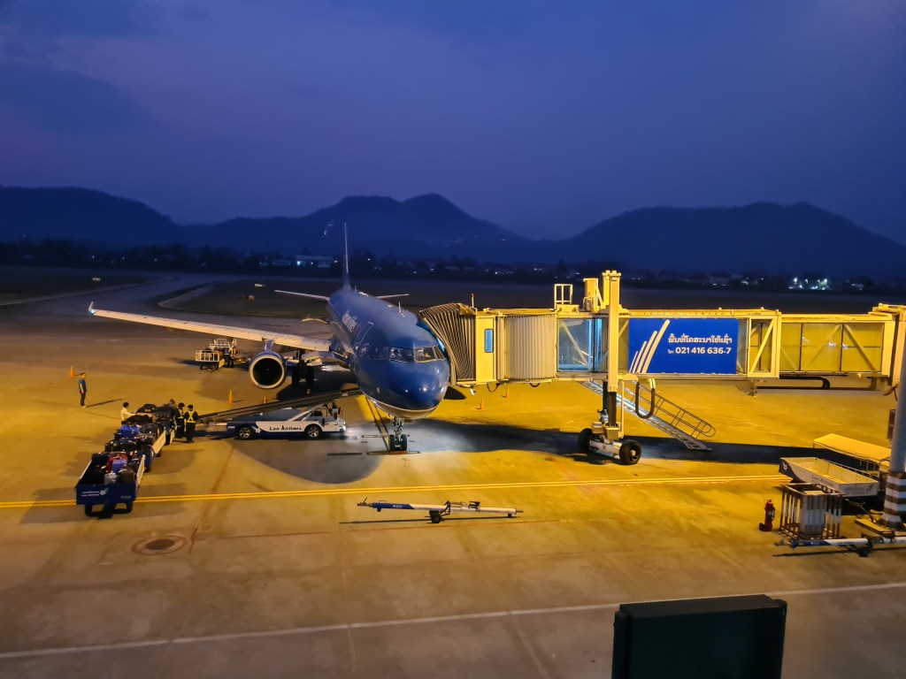 An airplane is parked at an airport gate during twilight, with ground crew loading baggage onto the aircraft. Mountains are visible in the background.
