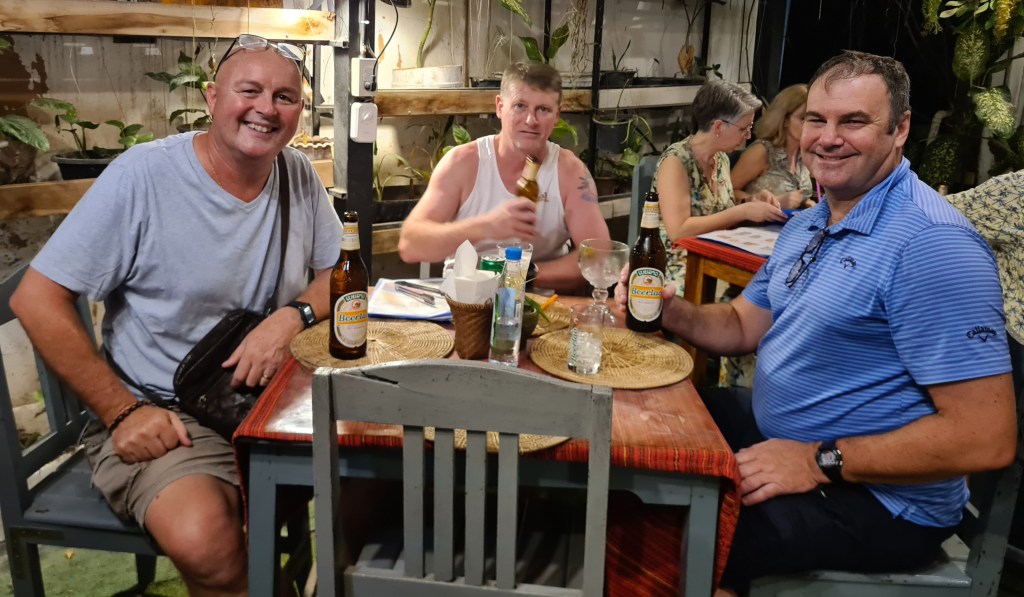 Three men sitting at a table in a restaurant, enjoying drinks and food. One man is smiling and holding a beer, another man is drinking from a glass, while the third is partially turned away. The setting includes plants and a casual dining atmosphere.