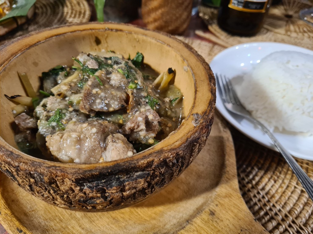 A bowl made from a coconut shell containing a savory meat and vegetable dish, garnished with herbs, accompanied by a plate of white rice.