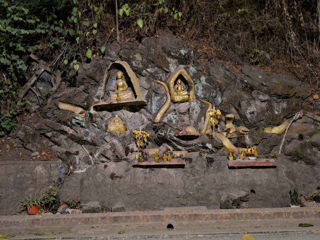 A rocky hillside featuring multiple golden sculptures and shrines, adorned with flowers and offerings.
