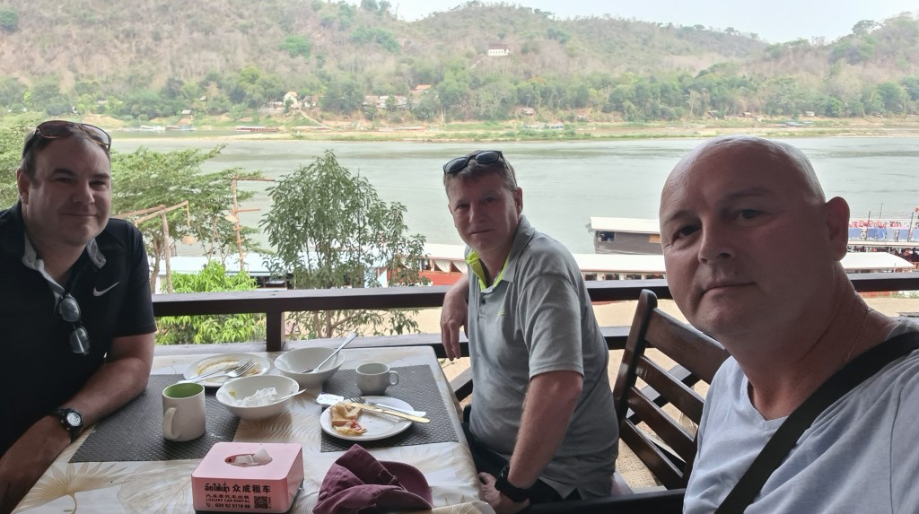 Three men sitting at a table with dishes and drinks, overlooking a river and mountains in the background.