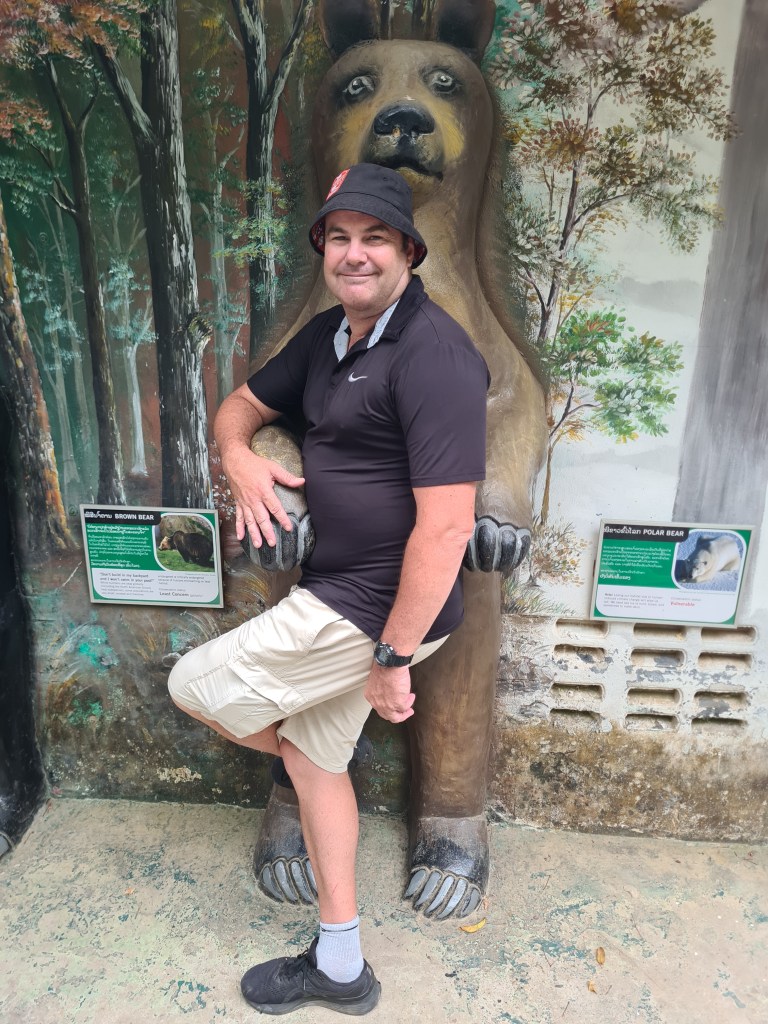 A man posing with a life-sized bear sculpture in a forest-themed setting, wearing a black shirt and beige shorts.