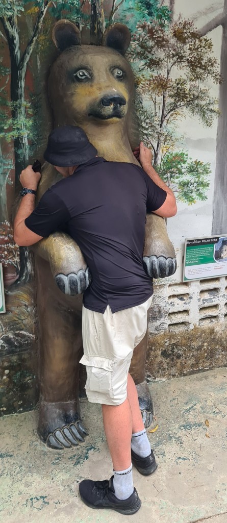 A person in a black shirt and shorts hugs a life-sized bear statue in front of a painted forest backdrop.